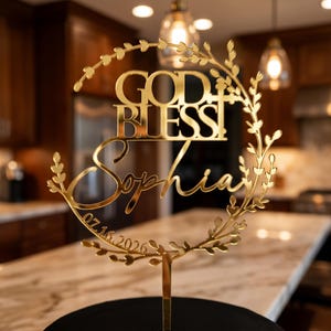Op de afbeelding: Een goudkleurige taarttopper met de woorden "GOD BLESS Sophia" en de datum "07.16.2026". De tekst staat in een cirkelvormige krans met bladeren. De topper staat op een zwarte basis.