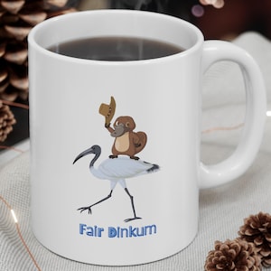 Könnte beinhalten: Eine weiße Keramik-Tasse mit schwarzem Henkel und dem Text "But First, Coffee" in fetter, schwarzer Schrift. Die Tasse steht auf einer Holzoberfläche. Die Tasse ist etwa 10 cm hoch.