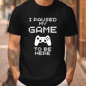 May include: Black t-shirt with the text "I PAUSED MY GAME TO BE HERE" in white, along with a white controller graphic. The shirt is a crew neck style.