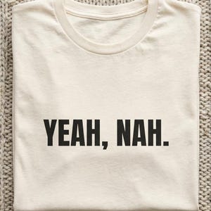 Könnte beinhalten: Ein gefaltetes, cremefarbenes T-Shirt mit dem Aufdruck "YEAH, NAH." in fetten, schwarzen Buchstaben. Das Shirt ist aus einem weichen Material gefertigt und wird auf einer strukturierten, neutral getönten Oberfläche präsentiert.