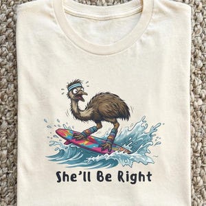May include: Cream-coloured t-shirt with a cartoon emu surfing on a colourful surfboard. The emu wears a headband and leg warmers. The text "She'll Be Right" is printed beneath the image.