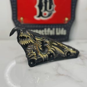 May include: A black and gold dragon-shaped helmet front piece featuring a detailed relief of a dragon with an open mouth and sharp teeth. Behind it sits a black and red shield-shaped plaque with the letter D and the text Respectful Lids.