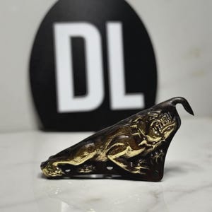 May include: A dark brown leather piece featuring a gold-toned embossed relief of a snarling panther in a pouncing position. The leather has visible stitching holes along the bottom edge. In the background, a black circular sign displays the letters DL.