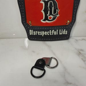 May include: A black and red leather firefighter helmet front piece featuring a central black letter D, four gold-colored rivets, and the text Disrespectful Lids in white. Below it lies a black leather strap with a silver-toned metal D-ring.