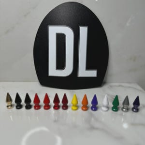 May include: A black, arch-shaped display sign featuring the white letters DL in the center. Arranged in a horizontal row in front of the sign are fourteen small, pointed, cone-shaped studs in various colors including gold, black, red, yellow, orange, purple, white, green, silver, and blue.