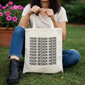 May include: A white canvas tote bag with black text that reads "YOUR DESIGN HERE" repeated ten times. The bag is being held by a person wearing blue jeans and a white t-shirt.