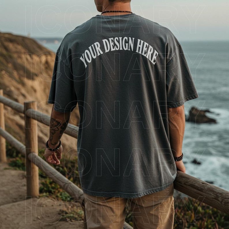 Puede incluir: Una persona lleva una camiseta gris oscura con el texto "YOUR DESIGN HERE" impreso en la espalda. La persona est&aacute; de pie en una pasarela de madera con vistas a una masa de agua.