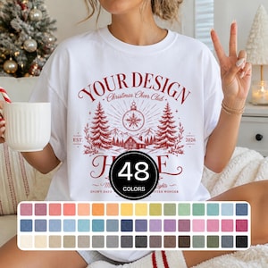 May include: A white crewneck t-shirt featuring a red graphic print with the text "YOUR DESIGN," "Christmas Cheer Club," "EST. 2026," and "SNOWY DAYS." The design includes pine trees, a cabin, and a snowflake ornament. A color chart shows 48 options.