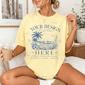 May include: A pale yellow t-shirt featuring a blue graphic print with the text "YOUR DESIGN Coastal Club HERE" and "EST. 2026". The design includes illustrations of palm trees, a sunset over the ocean, seashells, and a starfish.