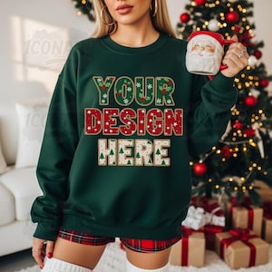 Puede incluir: Una sudadera navideña verde oscuro con el texto "YOUR DESIGN HERE" en rojo, verde y blanco. La sudadera se combina con pantalones cortos de cuadros rojos y una taza de Papá Noel. Un árbol de Navidad y regalos envueltos están en el fondo.
