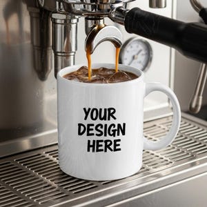 May include: A white coffee mug with the text "YOUR DESIGN HERE" in black, filled with iced coffee. The mug is positioned on a stainless steel espresso machine, with coffee pouring into the mug.