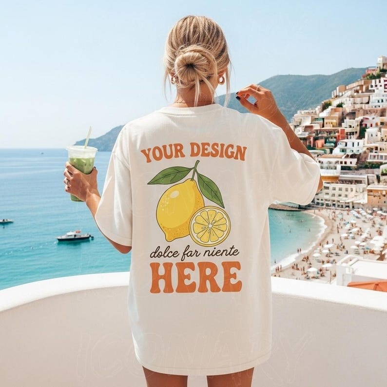 May include: Cream-colored t-shirt with a lemon graphic and the text "YOUR DESIGN dolce far niente HERE." The design features a whole lemon and a lemon slice with green leaves. The shirt is worn by a person holding a green drink.