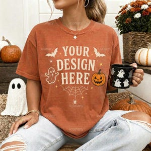 May include: A rust-colored short-sleeve t-shirt featuring the text "YOUR DESIGN HERE" in a cream-colored font. The graphic includes illustrations of a jack-o'-lantern, a spider web, bats, and ghosts. The shirt has a crew neckline and a relaxed fit.