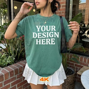 May include: A green t-shirt with the text "YOUR DESIGN HERE" in white. The shirt is paired with a white pleated skirt. The image also includes a black shoulder bag and a gold necklace.