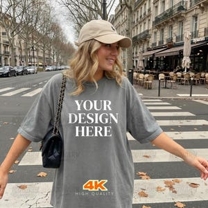 May include: Gray short-sleeve t-shirt with the text "YOUR DESIGN HERE" in white. The shirt also has a "4K HIGH QUALITY" logo. The person is wearing a beige baseball cap and carrying a black purse with a chain strap.