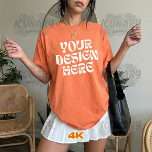 May include: Orange t-shirt with the text "YOUR DESIGN HERE" in white, paired with a white pleated skirt and a black handbag. The image also features a wicker chair and a mirror in the background.