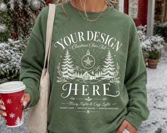 May include: An olive green crewneck sweatshirt featuring a white graphic print with the text YOUR DESIGN HERE, Christmas Cheer Club, Merry Nights & Cozy Lights, Snowy Days, Holiday Magic, and Winter Wonder, depicting a cabin, trees, and ornament.