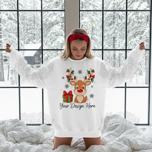 Könnte beinhalten: Weißes Sweatshirt mit einem Cartoon-Rentier-Design, das eine rote Nase, mit Stechpalmen verzierte Geweihe und eine Geschenkbox aufweist. Der Text "Your Design Here" ist unter dem Rentier aufgedruckt. Eine Person mit einem roten Stirnband trägt das Sweatshirt.