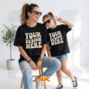Puede incluir: Una mujer y una ni&ntilde;a con camisetas negras con el texto "YOUR DESIGN HERE" en una fuente retro. Ambas llevan gafas de sol y vaqueros. La mujer est&aacute; sentada en un taburete de madera. La ni&ntilde;a est&aacute; de pie y hace la se&ntilde;al de la paz.
