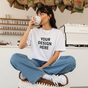 Puede incluir: Camiseta blanca con el texto "YOUR DESIGN HERE" en negro, combinada con vaqueros azules. La persona está sentada con las piernas cruzadas, sosteniendo una taza blanca. El fondo incluye sacos de café y estanterías.