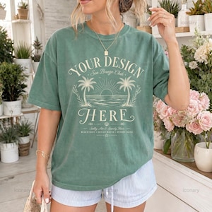 May include: A sage green short-sleeve t-shirt featuring a graphic print with the text "YOUR DESIGN HERE" above "Sea Breeze Club" and "Salty Air & Sandy Hair". The design includes illustrations of palm trees, a sun, and ocean waves.