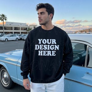 Könnte beinhalten: Schwarzes Sweatshirt mit Rundhalsausschnitt und dem weißen Text "YOUR DESIGN HERE". Das Sweatshirt wird von einer Person getragen, die neben einem hellblauen Oldtimer steht. Der Hintergrund zeigt einen Parkplatz und Palmen.