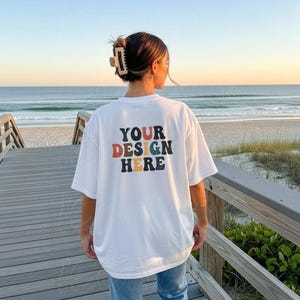 Op de afbeelding: Wit T-shirt met de tekst "YOUR DESIGN HERE" in retro-stijl letters. Het shirt wordt gedragen door een persoon op een houten promenade met uitzicht op een strand en de oceaan. De tekst is in een mix van kleuren: oranje, bruin, groen en geel.