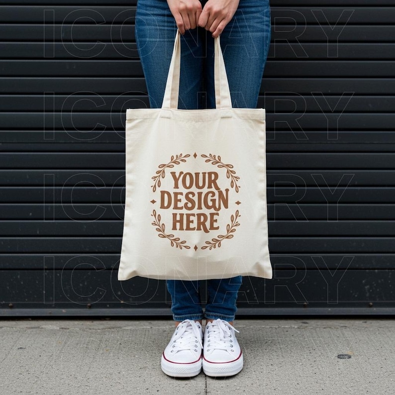 May include: A person is holding a white canvas tote bag with brown text that says "Your Design Here" surrounded by a wreath of leaves. The bag has brown straps and is in front of a black metal garage door.