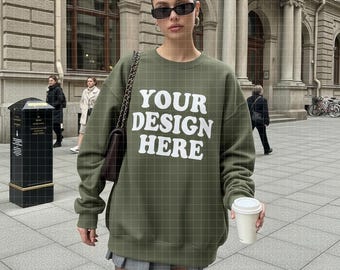 May include: An olive green sweatshirt with the text "YOUR DESIGN HERE" in white. The sweatshirt has a crew neck and long sleeves. The model is wearing sunglasses and holding a white coffee cup. A black purse with a chain strap is over the shoulder.