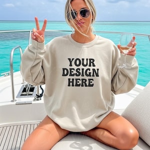 Puede incluir: Una sudadera beige con el texto "YOUR DESIGN HERE" en letras negras. La persona lleva gafas de sol y sostiene una copa de vino. El fondo es un océano turquesa.