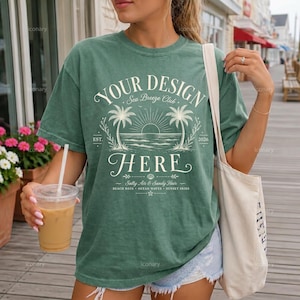 May include: A sage green short-sleeve t-shirt featuring a white graphic of palm trees, a sun, and ocean waves. The text reads: YOUR DESIGN HERE, Sea Breeze Club, EST. 2026, Salty Air & Sandy Hair, Beach Days, Ocean Waves, Sunset Skies.