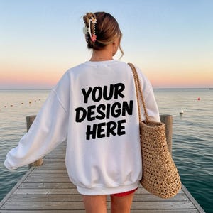 May include: White sweatshirt with the text "YOUR DESIGN HERE" in black. The person is wearing a red skirt and carrying a woven bag. The background shows a pier, the ocean, and a sunset.