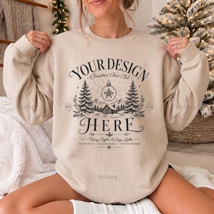 May include: A beige crewneck sweatshirt featuring a dark brown graphic print with the text "YOUR DESIGN HERE" above a winter scene with pine trees, a cabin, and a Christmas ornament. Additional text includes "Christmas Cheer Club", "EST. 2026", and "Merry Nights & Cozy Lights".