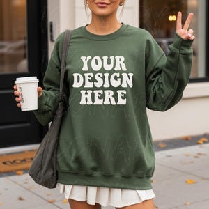 May include: An olive green sweatshirt with the text "YOUR DESIGN HERE" in white, a white coffee cup, and a gray shoulder bag. The person is giving a peace sign. The sweatshirt is a casual style.