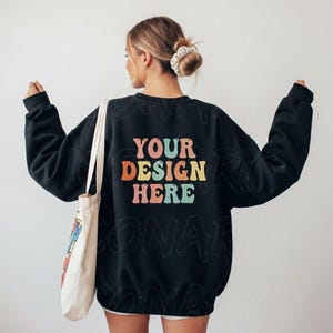 以下が含まれることがあります： カラフルなレトロフォントで「YOUR DESIGN HERE」と書かれた黒いクルーネックのスウェットシャツ。スウェットシャツは長袖でリラックスしたフィット感です。左側には白いトートバッグが見えます。