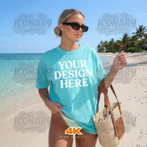 Puede incluir: Una camiseta turquesa con el texto "YOUR DESIGN HERE" en blanco, usada por una persona en una playa. La persona lleva gafas de sol y sostiene un bolso de mano de paja con detalles de cuero. El fondo presenta una playa, el océano y palmeras.