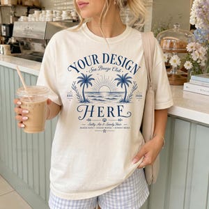 Può includere: Una t-shirt a maniche corte color bianco sporco con una grafica blu scuro di un tramonto sull'oceano con palme. Il testo recita: YOUR DESIGN HERE, Sea Breeze Club, EST. 2026, Salty Air & Sandy Hair, BEACH DAYS, OCEAN WAVES, SUNSET SKIES.