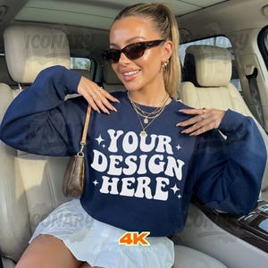 May include: Navy blue sweatshirt with the text "YOUR DESIGN HERE" in white. The sweatshirt is paired with a white skirt. The image also shows a small brown purse, sunglasses, and gold jewelry.