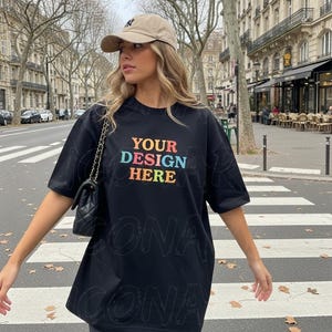 以下が含まれることがあります： カラフルな文字で「YOUR DESIGN HERE」と書かれた黒のオーバーサイズTシャツ。クルーネックと半袖です。ベージュの野球帽とチェーンストラップ付きの黒いハンドバッグも写っています。