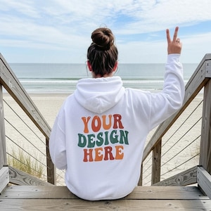 以下が含まれることがあります： オレンジ、緑、赤で「YOUR DESIGN HERE」の文字が入った白いパーカー。 海を背景に、ビーチへと続く木製の階段で着用されています。 その人はピースサインをしています。