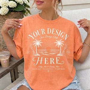 May include: An orange short-sleeve t-shirt featuring a white graphic print with the text YOUR DESIGN HERE, Sea Breeze Club, EST. 2026, and Salty Air & Sandy Hair. The design includes illustrations of two palm trees, a sun, and ocean waves.