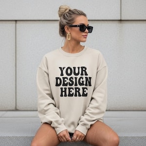 May include: A cream-coloured sweatshirt featuring the text "YOUR DESIGN HERE" in black, retro lettering. The long-sleeved sweatshirt has a crew neck. The model is wearing sunglasses and gold earrings.