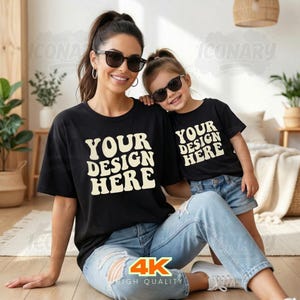 Könnte beinhalten: Eine Frau und ein Kind tragen passende schwarze T-Shirts mit dem Text "YOUR DESIGN HERE" in einer Retro-Schriftart. Beide tragen Sonnenbrillen und blaue Jeans. Die Frau sitzt auf dem Boden, und das Kind lehnt sich an ihre Schulter.