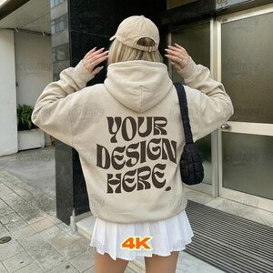 Op de afbeelding: Een beige hoodie met de tekst "YOUR DESIGN HERE" in bruine letters op de rug. De persoon draagt een beige baseballpet en een witte geplooide rok. Een zwarte schoudertas is zichtbaar.