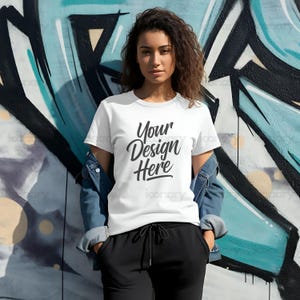 Może przedstawiać: Biała koszulka z napisem "Your Design Here" czarnymi literami. Koszulkę nosi osoba stojąca przed ścianą z graffiti.