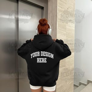 Op de afbeelding: Zwarte hoodie met de tekst "YOUR DESIGN HERE" in witte blokletters op de rug. De persoon draagt een witte korte broek en een koptelefoon. De achtergrond omvat een zilveren liftdeur en een beige muur.