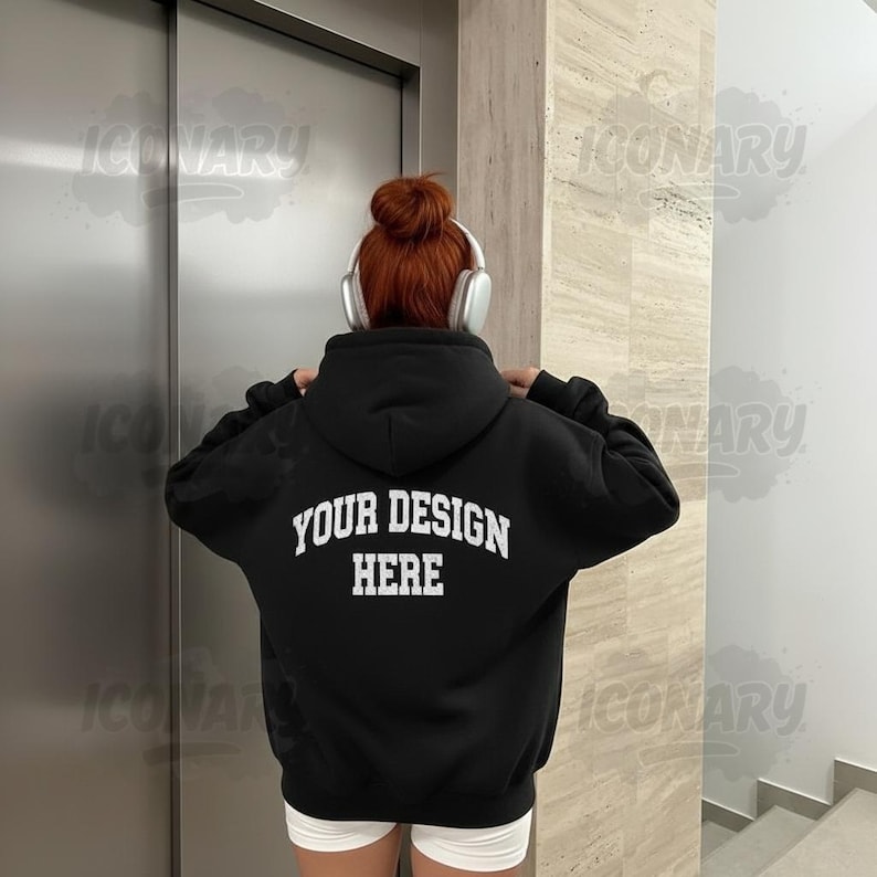 Op de afbeelding: Zwarte hoodie met de tekst "YOUR DESIGN HERE" in witte blokletters op de rug. De persoon draagt een witte korte broek en een koptelefoon. De achtergrond omvat een zilveren liftdeur en een beige muur.