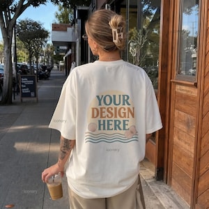 May include: Cream-colored t-shirt with the text "YOUR DESIGN HERE" in a retro design. The design includes a half-circle, seashells, and wavy lines. The person is holding a coffee drink in a clear plastic cup.