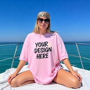 以下が含まれることがあります： 黒い文字で「YOUR DESIGN HERE」と書かれた、明るいピンク色のオーバーサイズのTシャツ。サングラスとレオパード柄のバンダナを着用。背景には、澄み切った青い空と海が広がっています。