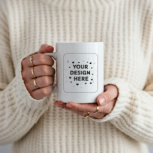 Pode incluir: Caneca de cerâmica branca com um design preto que diz "YOUR DESIGN HERE" rodeado de pequenos corações. A caneca está sendo segurada por uma pessoa que está vestindo um suéter branco.